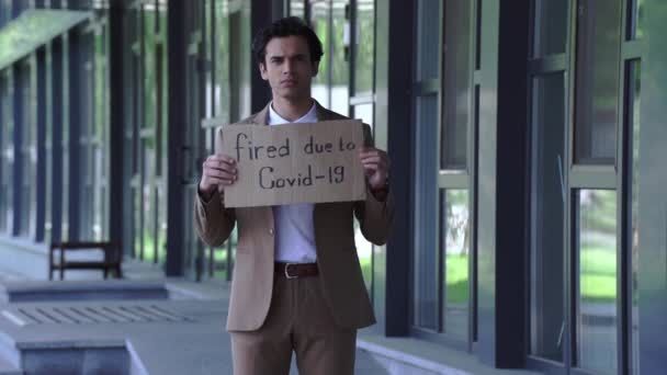 triste manager in possesso di cartone con licenziato a causa di covid-19 lettering vicino edificio - Filmati, video