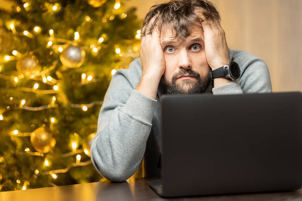 un hombre cuyo cansado shaggy se sienta en un ordenador portátil en la noche de Navidad - Foto, imagen