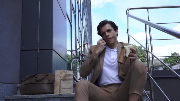 kiireinen liikemies puhuu kännykkään syödessään voileipä ulkopuolella - Materiaali, video