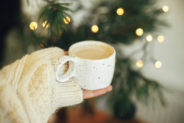 Hand in cozy sweater holding warm coffee in stylish cup on background of fir branches with warm lights in festive scandinavian room. Atmospheric winter time and hygge home. Merry Christmas! - Photo, image