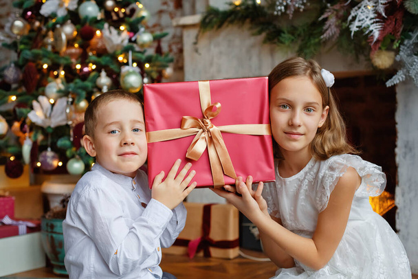 Τα παιδιά έσκυψαν τα κεφάλια τους στο δώρο. Ένα κορίτσι και ένα αγόρι με δώρα κάτω από το χριστουγεννιάτικο δέντρο. Αδελφός και αδελφή είναι ευχαριστημένοι με τα δώρα της Πρωτοχρονιάς. - Φωτογραφία, εικόνα