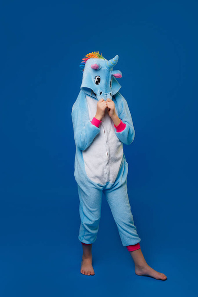 Une fille en costume de licorne bleu confortable en pleine hauteur, couvrant son visage avec une capuche, pose dans le studio sur un fond bleu isolé. Place pour le texte - Photo, image