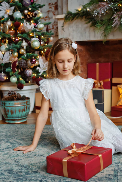 A girl in a white dress with a red gift box in a room decorated for Christmas. Sitting by the fireplace and the Christmas tree, looking at the gift and pulling the ribbon. - Foto, imagen