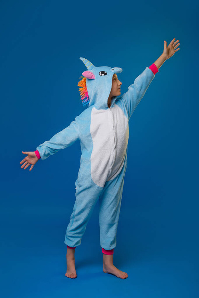 A girl in a cozy blue unicorn costume in full height, with her arms outstretched to the sides, poses in the studio on a blue isolated background. Place for text - Photo, Image