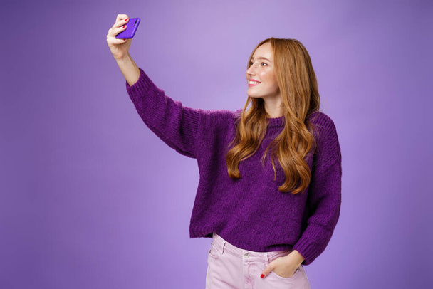 Attractive confident and stylish happy 20s woman with ginger hair standing carefree with joyful smile over purple background extending hand with smartphone as taking selfie happily - Фото, зображення