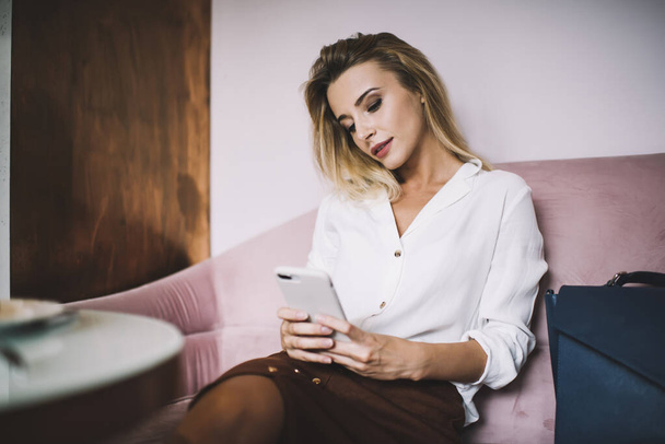 Thoughtful young female in white shirt and brown skirt checking notifications on phone while sitting on couch near table in light cafe - Фото, изображение