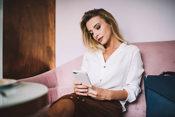 Przemyślana młoda kobieta w białej koszuli i brązowej spódnicy sprawdza powiadomienia przez telefon siedząc na kanapie przy stole w jasnej kawiarni - Zdjęcie, obraz