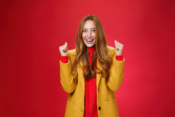 Ура успеху. Портрет счастливой веселой и искренней милой рыжей женщины в желтом пальто, поднимающей сжатые кулаки и празднующей радостные позитивные новости, торжествующей над победой и широко улыбающейся - Фото, изображение