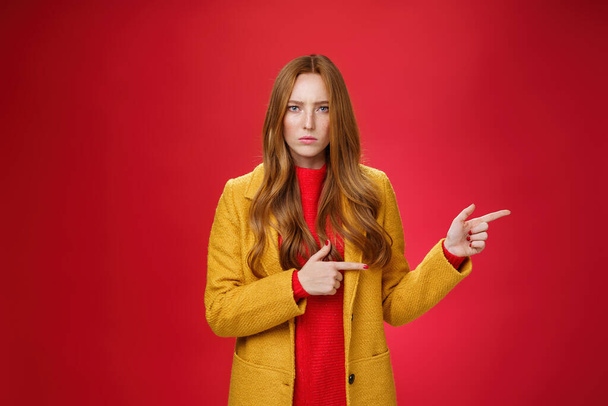 Dühös és sértett csalódott vörös hajú nő sárga köpenyben ráncolja szigorú és komoly a kamera előtt ellenszenv és agresszió rámutatva jobbra kér magyarázatot magad felett piros háttér - Fotó, kép