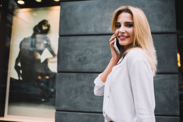 Side view of confident young female with blond hair wearing talking via mobile phone while walking against facade of urban building and looking at camera - Фото, зображення