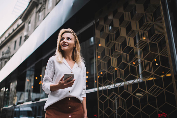 Low angle of cheerful young female in trendy outfit walking on street beside modern building and browsing mobile phone looking away - Фото, зображення