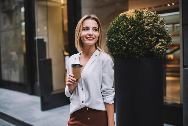 Cheerful young female in white blouse standing near shop entrance with lush green bush and drinking coffee to go looking away - Foto, afbeelding