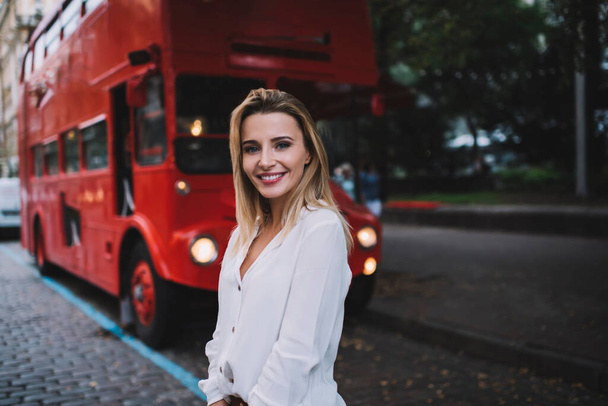 Joven turista sonriente con el pelo largo rubio en camisa blanca de pie cerca de autobús turístico en la calle y mirando a la cámara - Foto, imagen