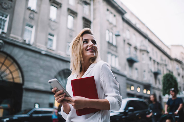 From below of young female entrepreneur in elegant outfit smiling and looking away while standing on street and browsing smartphone - Fotografie, Obrázek