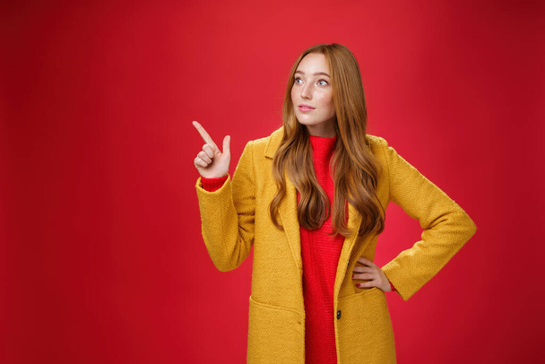 Wondered curious good-looking redhead woman in coat stopping on way looking and pointing at upper left corner curious and interested, observing surprising promotion over red background - Foto, Imagen