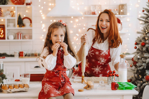 A little girl in the New Year's kitchen is sitting on the table with cookies in her hands, and her mother is preparing dough. - Valokuva, kuva