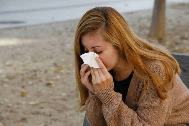 Sick woman sneezing and blowing her nose in a handkerchief tissue, outdoor in a park - Φωτογραφία, εικόνα