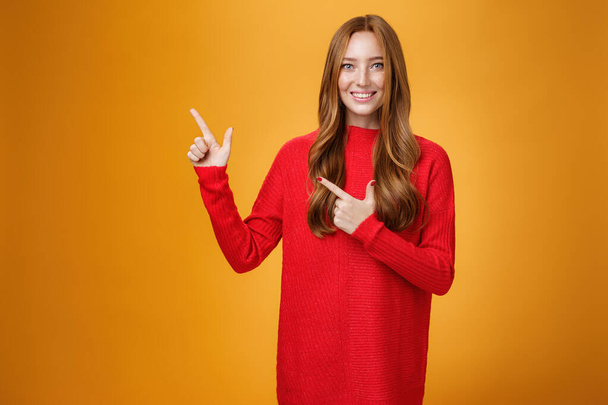 Assertive and helpful charming young female assitant in red knitted dress with ginger hair smiling friendly and pleasant as pointing at upper left corner, showing interesting thing over orange wall - Foto, afbeelding