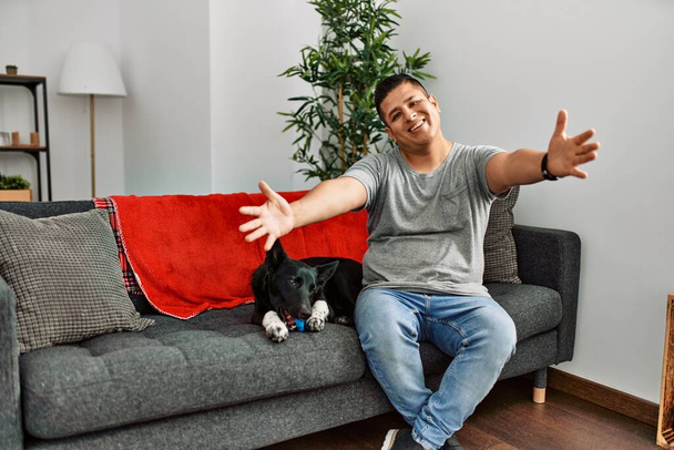 Junger lateinischer Mann und Hund, die zu Hause auf dem Sofa sitzen und mit offenen Armen in die Kamera lächeln, umarmen sich. fröhlicher Ausdruck, der das Glück umarmt.  - Foto, Bild