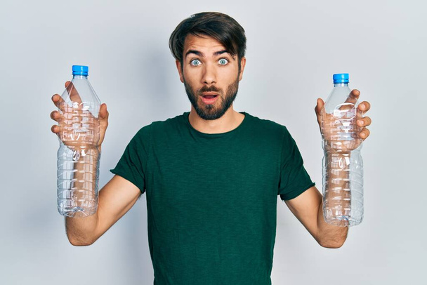 Joven hombre hispano sosteniendo botellas de plástico de reciclaje asustado y sorprendido con la expresión sorpresa y asombrado, miedo y cara emocionada.  - Foto, Imagen