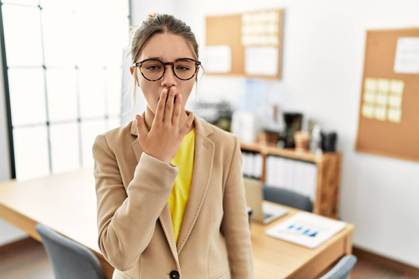 Νεαρή μελαχρινή έφηβη που φοράει επιχειρηματικό στυλ στο γραφείο βαριέται χασμουρητό κουρασμένο να καλύπτει το στόμα με το χέρι. ανησυχία και υπνηλία.  - Φωτογραφία, εικόνα