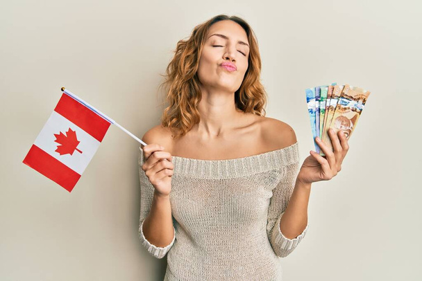 Młoda kaukaska kobieta trzymająca flagę Kanady i dolary patrząca w kamerę i dmuchająca pocałunek jest urocza i seksowna. wyrażenie miłości.  - Zdjęcie, obraz