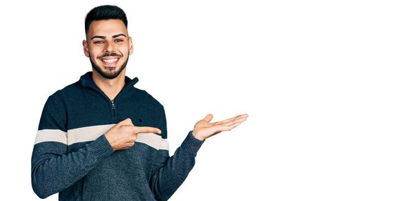Νεαρός Ισπανόφωνος με γενειάδα φορώντας casual χειμερινό πουλόβερ κατάπληκτος και χαμογελώντας στην κάμερα ενώ παρουσιάζει με το χέρι και δείχνοντας με το δάχτυλο.  - Φωτογραφία, εικόνα