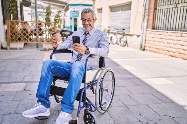Moyen Age homme hispanique assis sur fauteuil roulant en utilisant un smartphone dans la rue - Photo, image