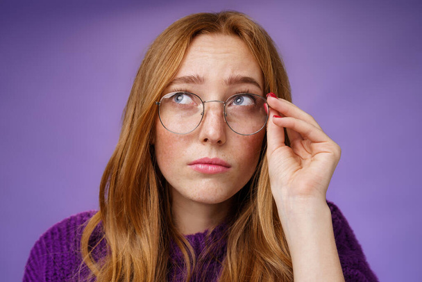 Clumsy and cute young female teacher in glasses with red hair looking unsure and confused looking interested at upper left corner touching rim of glasses thinking or picturing in imagination - Foto, afbeelding