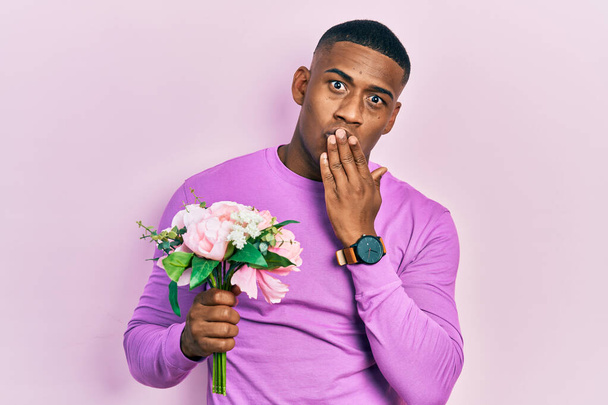 Młody czarny mężczyzna trzyma bukiet białych kwiatów okrywających usta dłonią, zszokowany i przerażony pomyłką. zaskoczona ekspresja  - Zdjęcie, obraz