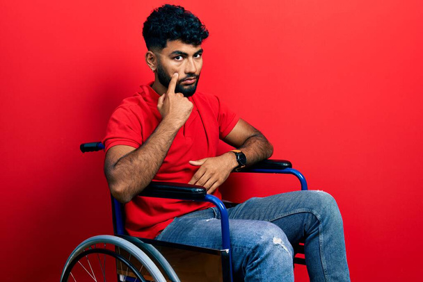 Arabský muž s vousy sedící na kolečkovém křesle ukazující na oko a pozorující tvé gesto, podezřívavý výraz  - Fotografie, Obrázek