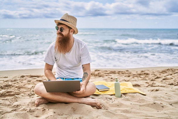 Fiatal vörös hajú férfi laptoppal ül a törölközőn a strandon. - Fotó, kép