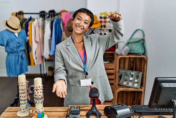 Młoda Latynoska kobieta z krótkimi włosami pracująca jako menedżer w detalicznym butiku, patrząca w kamerę uśmiechnięta z otwartymi ramionami na uścisk. radosna ekspresja obejmująca szczęście.  - Zdjęcie, obraz