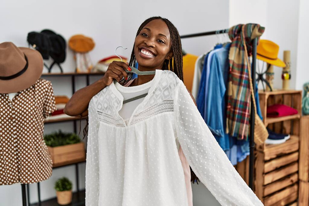 Νεαρή Αφρο-Αμερικανίδα πελάτισσα χαμογελά χαρούμενη κρατώντας κρεμάστρα με ρούχα στο κατάστημα ρούχων. - Φωτογραφία, εικόνα