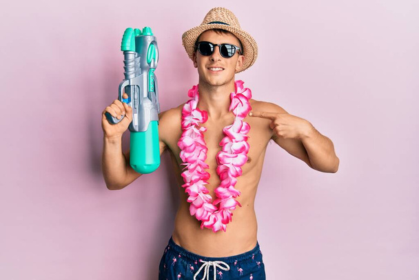 Young caucasian man wearing swimsuit and hawaiian lei holding watergun pointing finger to one self smiling happy and proud  - Fotó, kép