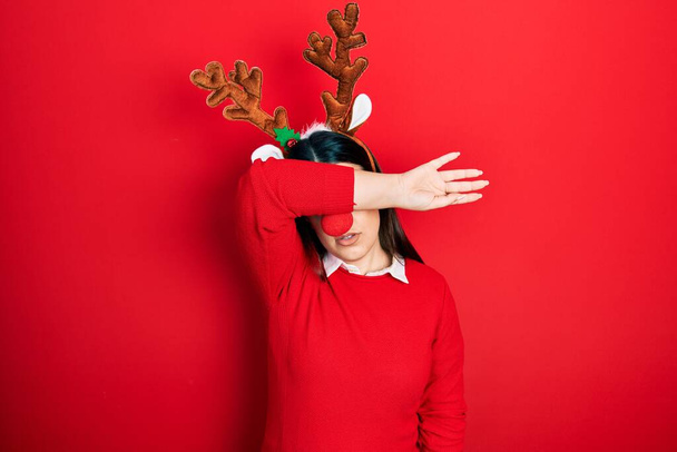 Young hispanic woman wearing deer christmas hat and red nose covering eyes with arm, looking serious and sad. sightless, hiding and rejection concept  - Photo, Image