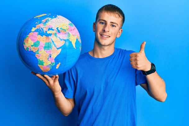 Young caucasian man holding world ball smiling happy and positive, thumb up doing excellent and approval sign  - Zdjęcie, obraz