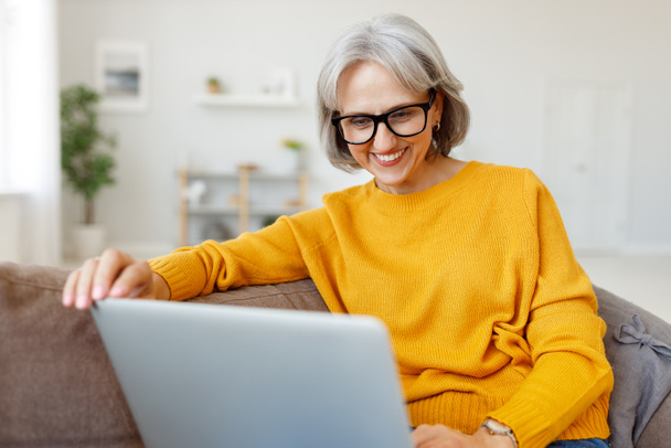 Portrait of happy smiling senior woman resting relaxing on sofa with laptop at home - Photo, Image