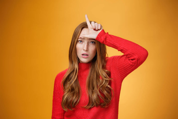 Arrogant and self-satisfied redhead woman humiliating person showing loser sign on forehead mocking and disdain rival standing confident and snobbish over orange background - Photo, Image