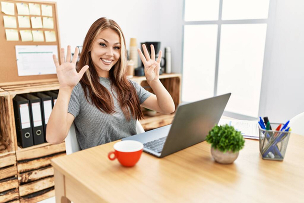 Νεαρή μελαχρινή γυναίκα που εργάζεται στο γραφείο με φορητό υπολογιστή που δείχνει και δείχνει προς τα πάνω με τα δάχτυλα νούμερο εννέα, ενώ χαμογελά αυτοπεποίθηση και χαρούμενος.  - Φωτογραφία, εικόνα