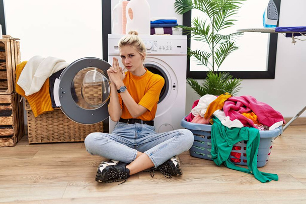 Junge blonde Frau, die Wäsche wäscht, sitzt an der Waschmaschine und hält symbolische Pistole mit Handgeste, spielt Tötungswaffen, wütendes Gesicht  - Foto, Bild