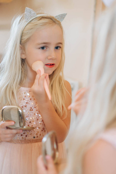 Linda niña jugando con cosméticos mirándose en el espejo. Niña sosteniendo polvo maquillaje cepillo - Foto, Imagen