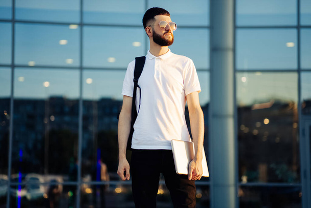 young male student in sunglasses with a backpack and a laptop, stands on the street of the city, looks to the side thoughtfully. Young man near building outdoors - Фото, зображення