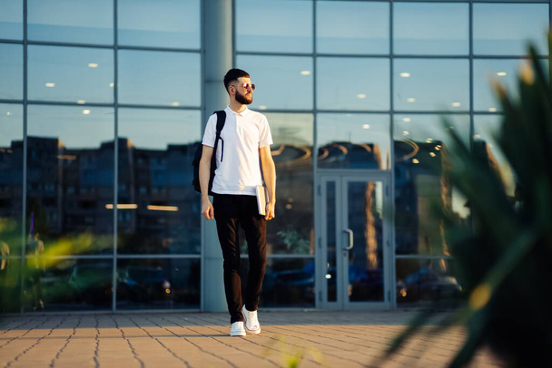 A student walking around the city with a backpack and a laptop. Attractive young man in sunglasses walks down the street on a sunny day, education concept - Zdjęcie, obraz