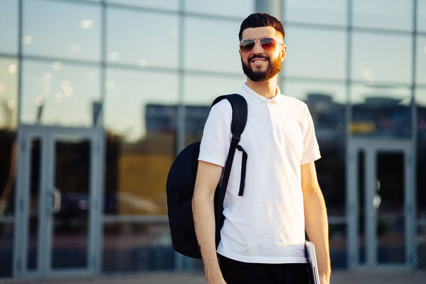 young male student in sunglasses with a backpack and a laptop, stands on the street of the city, looks to the side thoughtfully. Young man near building outdoors - Fotoğraf, Görsel