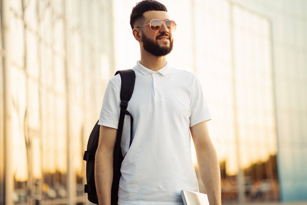 Business man gathering in sunglasses and with a black backpack walking along a city street with a laptop in his hands, a successful man in casual clothes walking outdoors near a building - 写真・画像