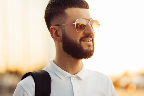 handsome man with a beard in sunglasses, in a casual white shirt on a sunny day, smiles. Man outdoors near a glass building, Fashion man, on a street background - Fotografie, Obrázek
