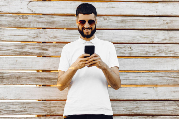 Handsome young bearded man using his phone with a smile while standing on a wooden background outdoors, happy man with glasses typing sms on smartphone - Foto, imagen