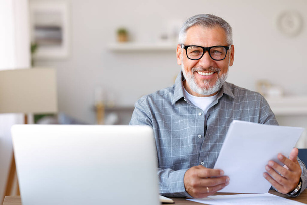 . Красивый улыбающийся пожилой человек читает финансовые документы, работая удаленно на ноутбуке дома - Фото, изображение