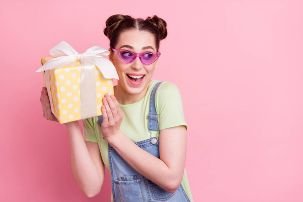 Photo of crazy girl hold present box look empty space wear sunglass jeans overall green t-shirt isolated on pink background - Foto, immagini